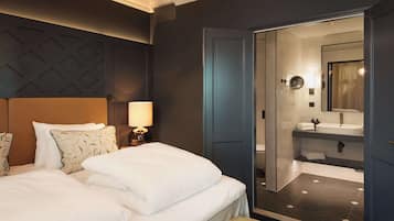 Una habitación de hotel con una cama, una mesita de noche con una lámpara, un baño con lavamanos y espejo, y una puerta que da al baño.