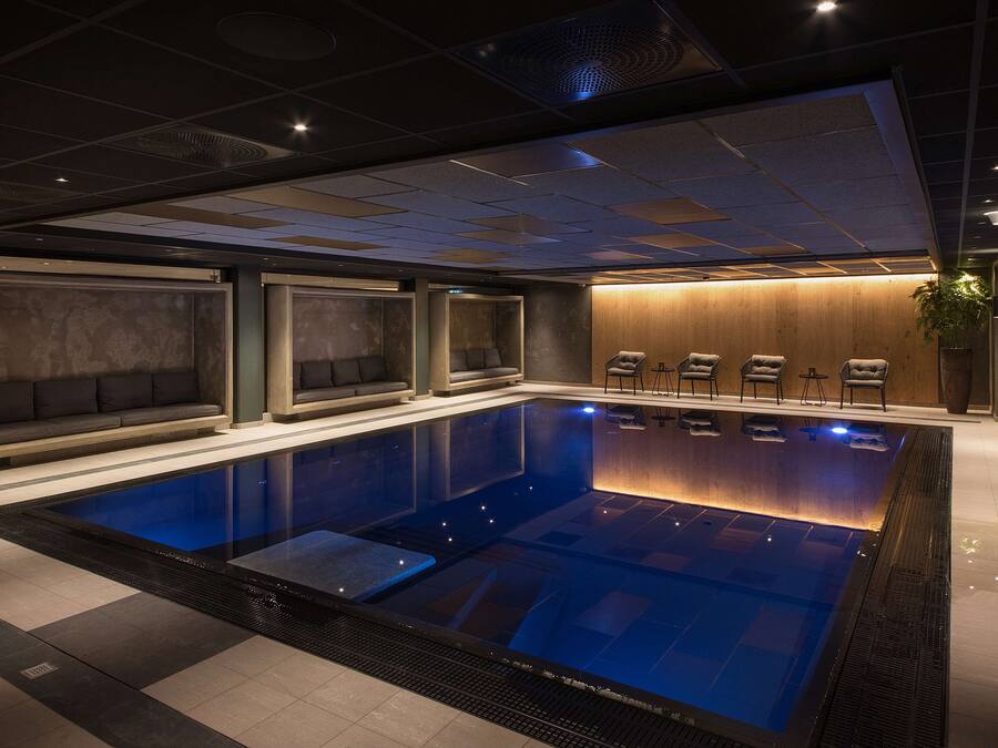 A modern indoor swimming pool with a clear water surface, surrounded by seating areas and a wooden wall.