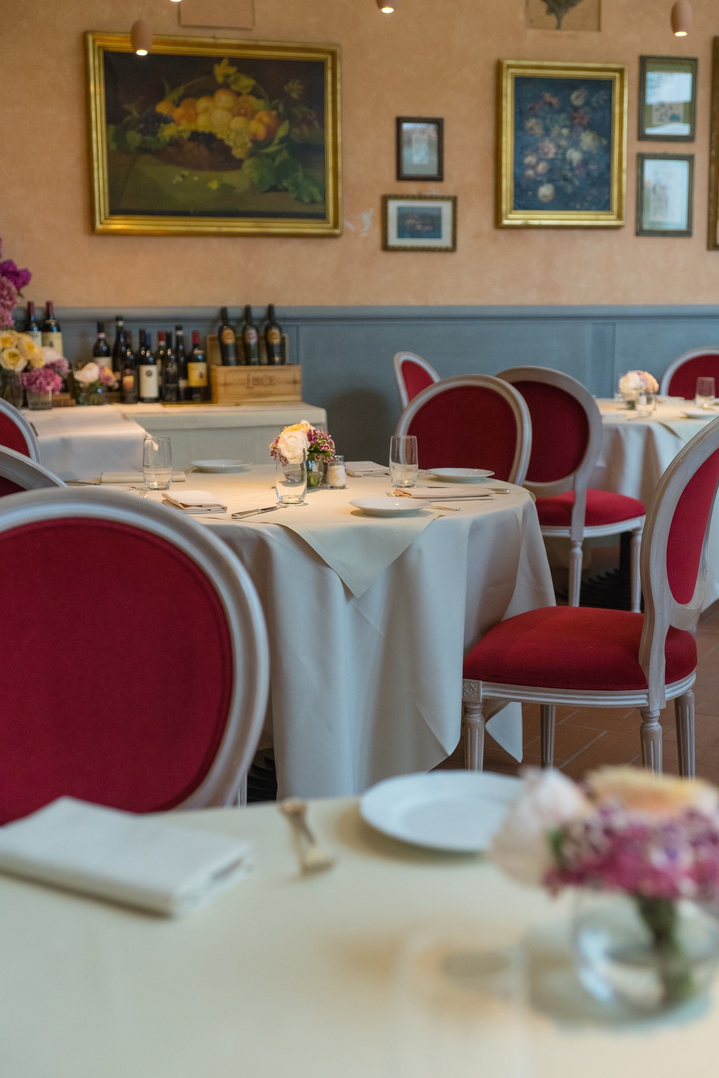 A restaurant interior with round tables, red upholstered chairs, white tablecloths, and framed floral paintings on the wall.
