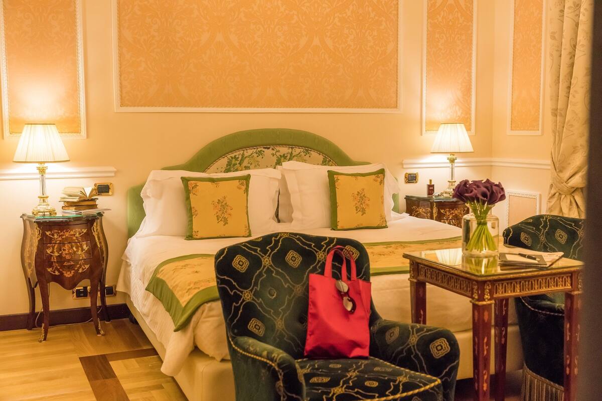 Ein Hotelzimmer mit einem Bett, zwei Nachttischen, einem Sessel, einem kleinen Tisch und einer Vase mit Blumen.