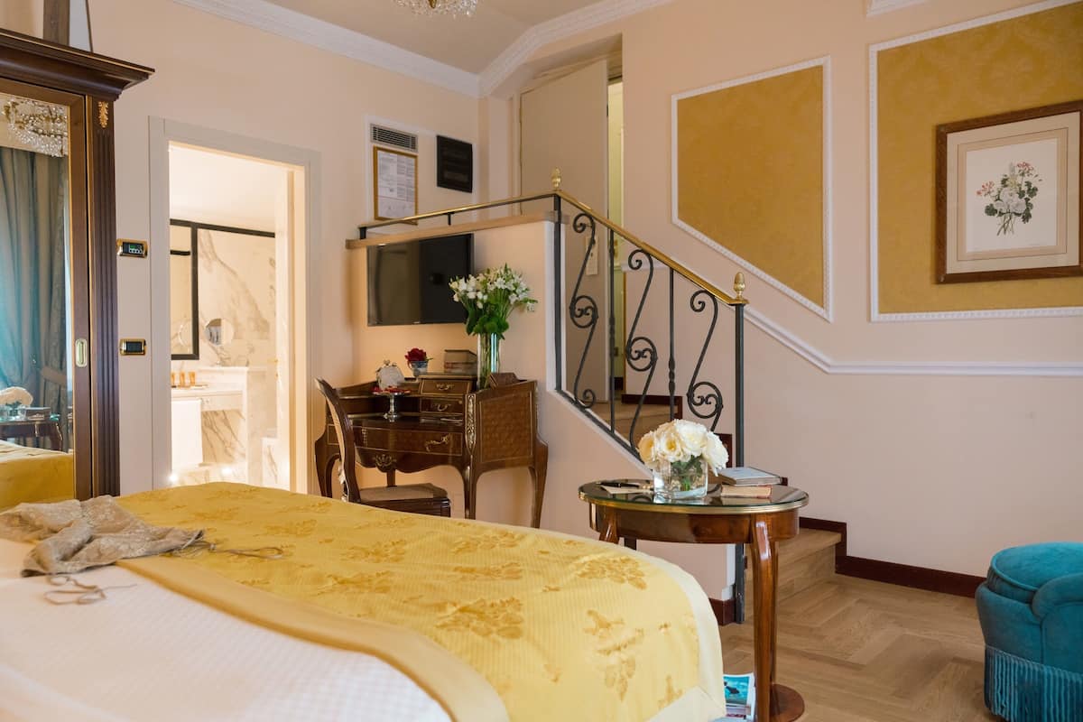 Ein Hotelzimmer mit einem Bett, einem Schreibtisch, einem Sessel, einem kleinen Tisch mit Blumen und einer Treppe.