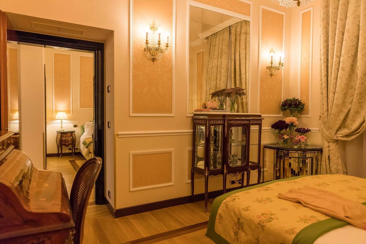 Ein Hotelzimmer mit einem Bett, einem Holzstuhl, einem kleinen Tisch mit Lampe, einem Spiegel und einer Kommode mit Blumen in einer Vase.