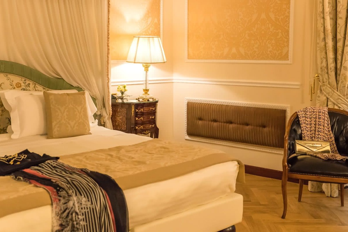 A hotel room with a bed, a chair, a nightstand, a lamp, and a radiator.