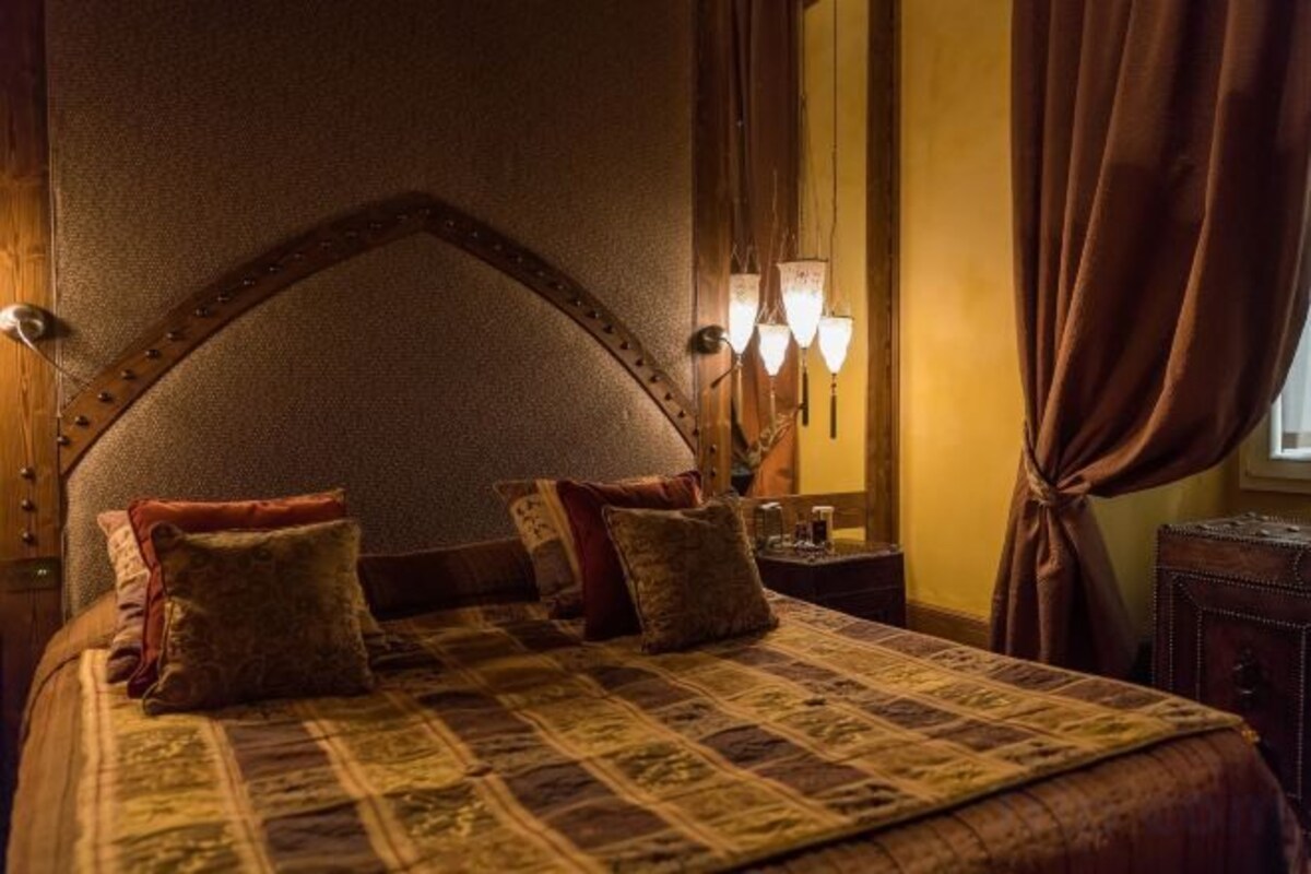 A bed with a headboard, pillows, and a quilt. There is a nightstand with a lamp and a mirror. The room has curtains and a window.