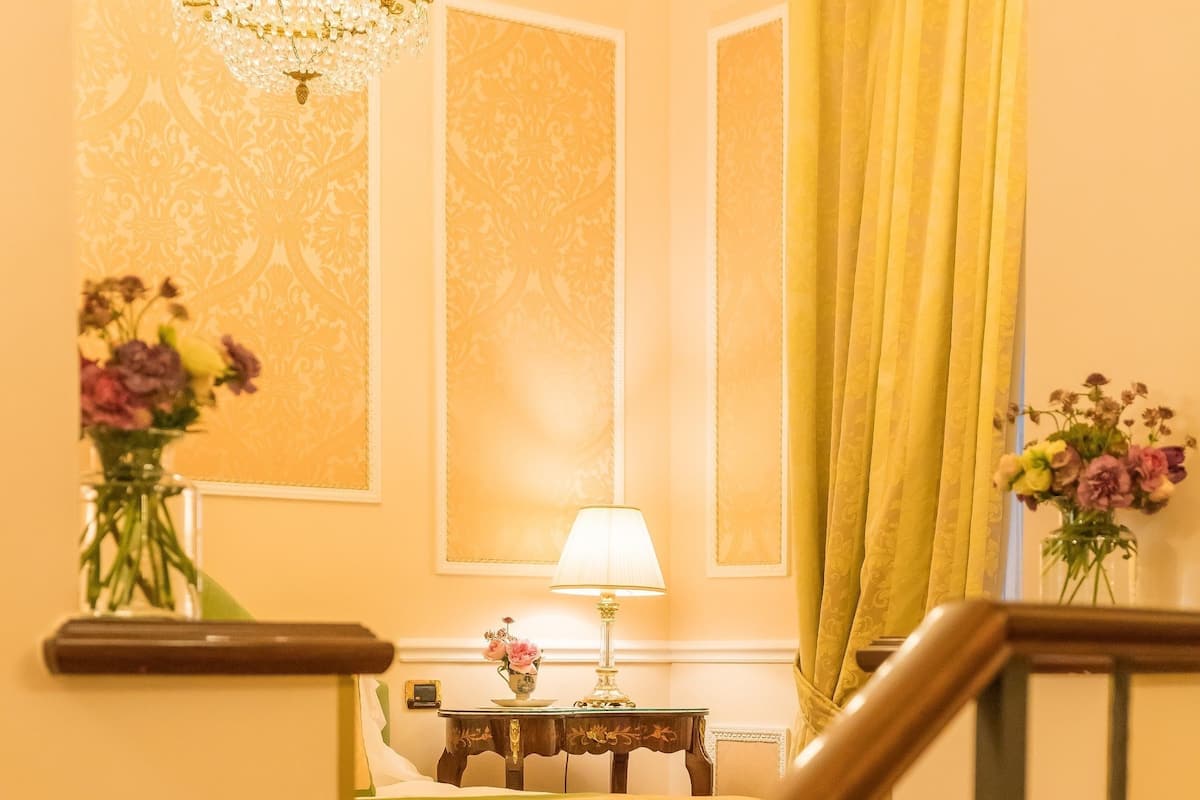 Ein Hotelzimmer mit Treppe, einem Chandelier, einem Bett mit grüner Bettwäsche, einem Nachttisch mit Lampe sowie zwei Vasen mit Blumen.