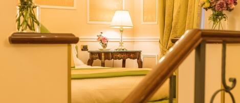 A hotel room with a staircase, a chandelier, a bed with green bedding, a nightstand with a lamp, and two vases with flowers.