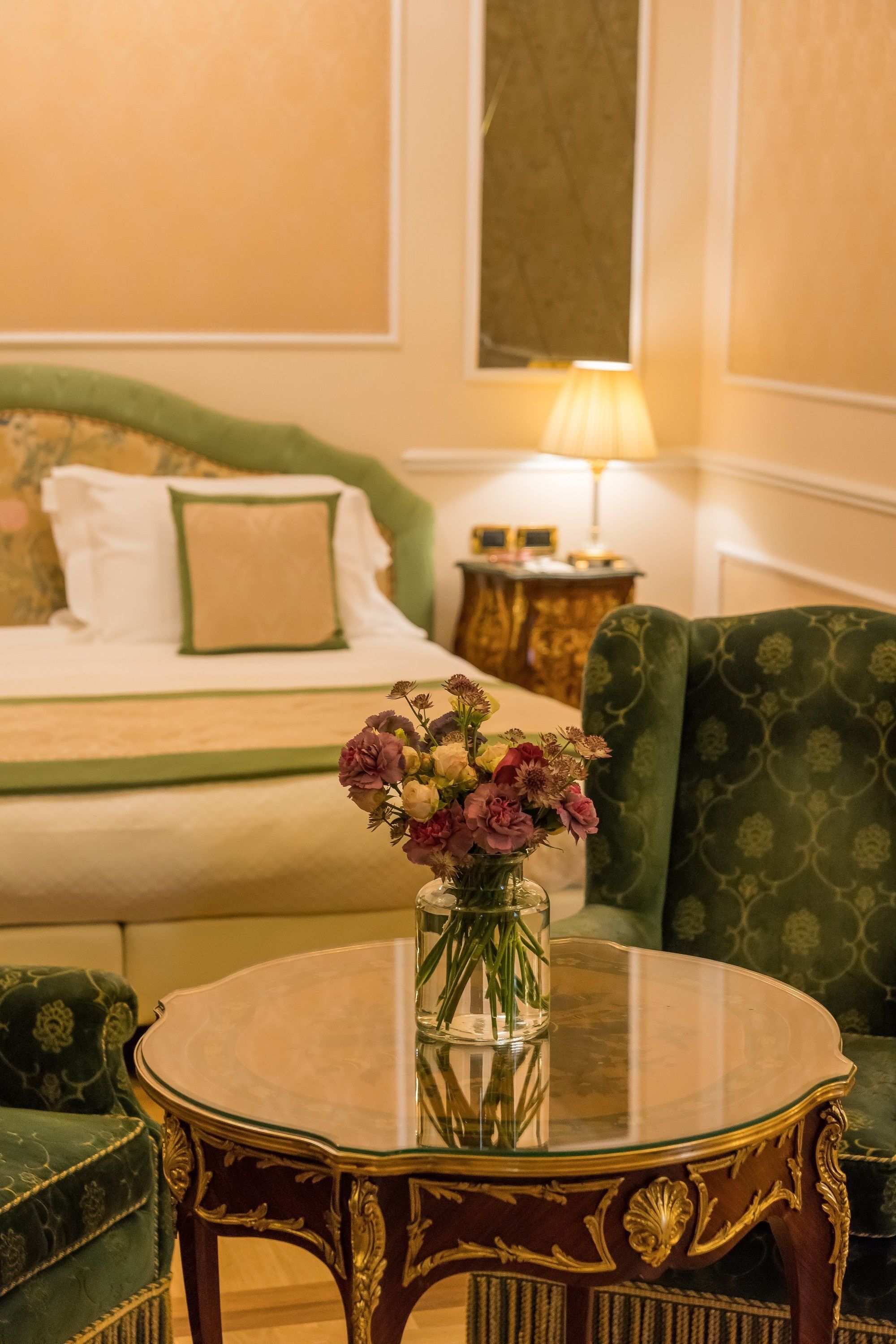 Habitación de hotel con cama, un sillón verde, una mesa redonda con un florero y una lámpara en una mesita auxiliar.