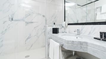 Un baño moderno con cabina de ducha de vidrio, pared con azulejos de mármol, lavamanos blanco y dispensador de jabón negro, y un espejo sobre el lavamanos.