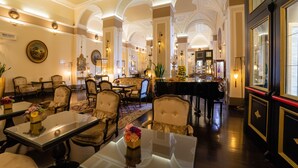 A grand lobby with high ceilings, ornate columns, and a grand piano. Tables and chairs are arranged for guests.