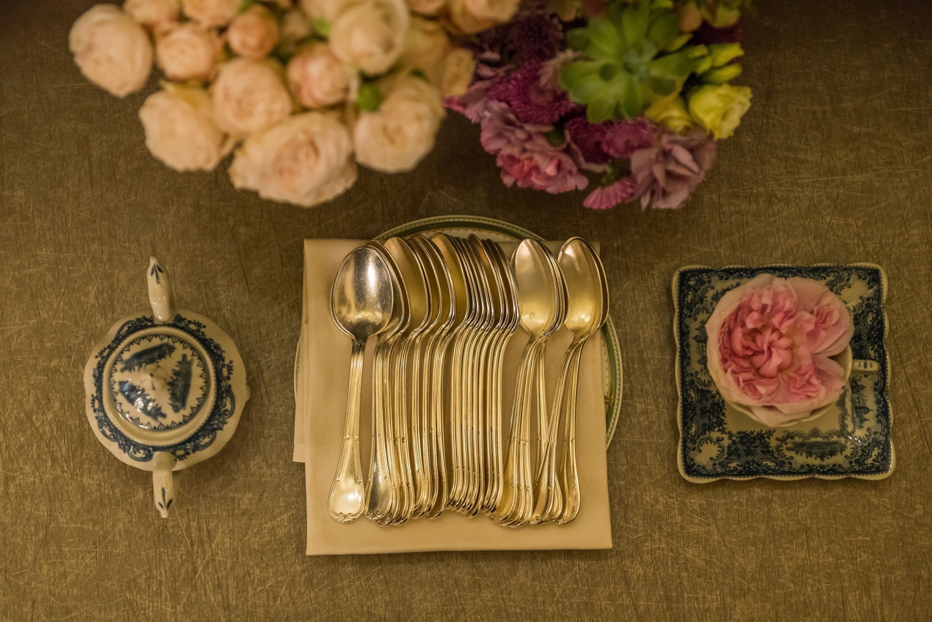 A set of gold spoons and a teapot on a beige napkin, with a blue and white plate with a pink flower beside them.