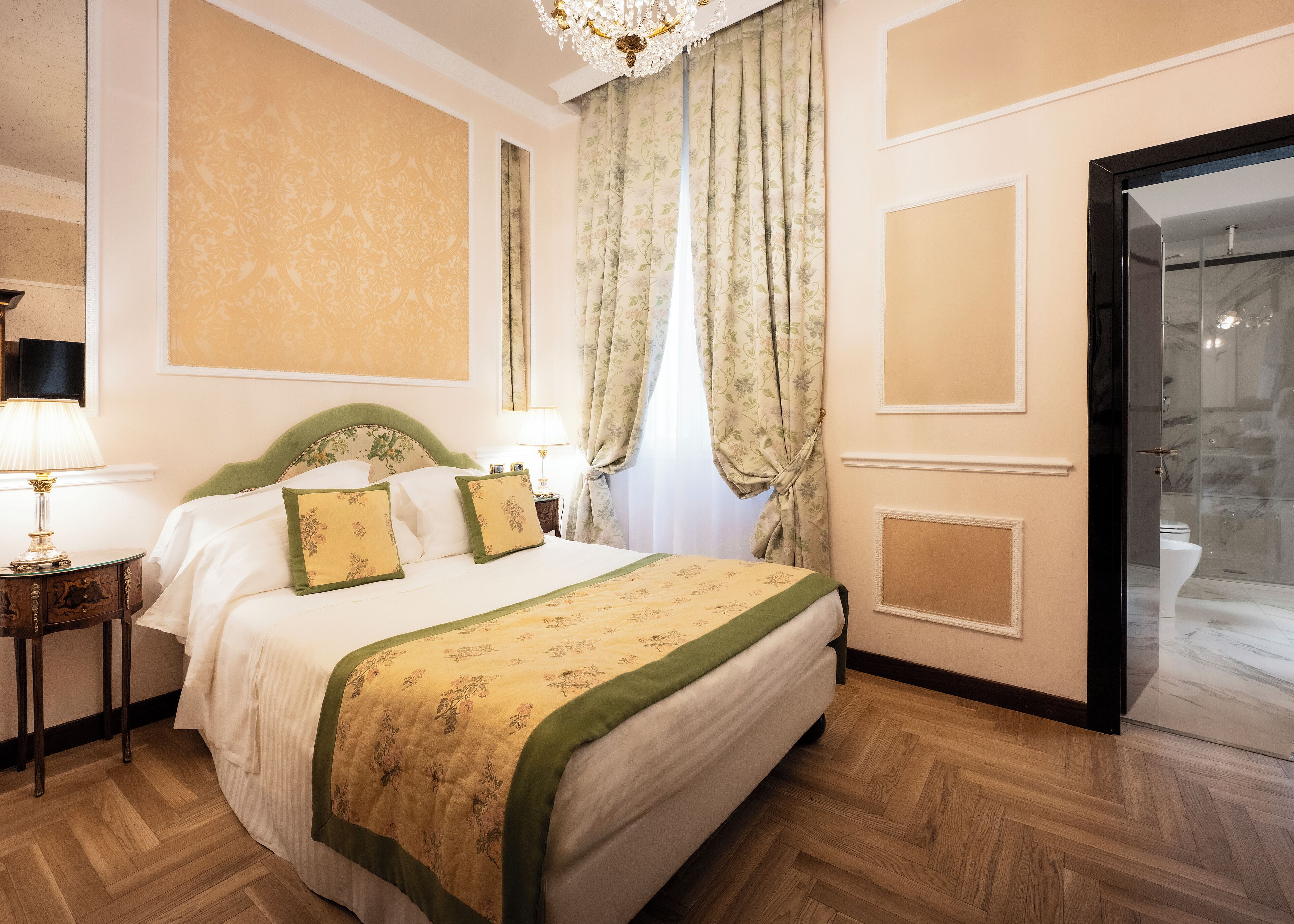 A hotel room with a large bed, a nightstand with a lamp, a mirror, and a view of the bathroom through a glass door.
