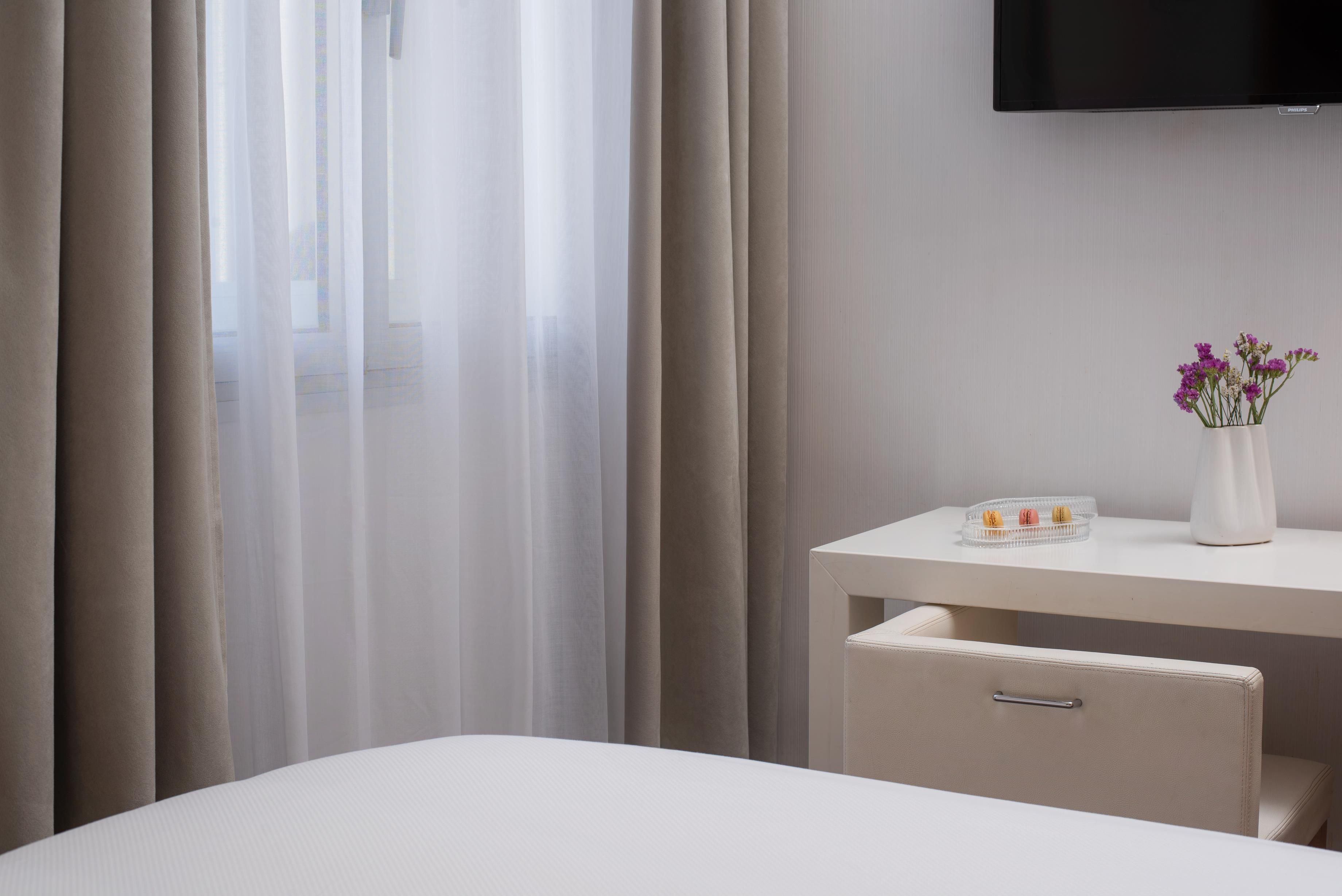 A hotel room with a bed, a desk with a vase of flowers, and a television mounted on the wall.