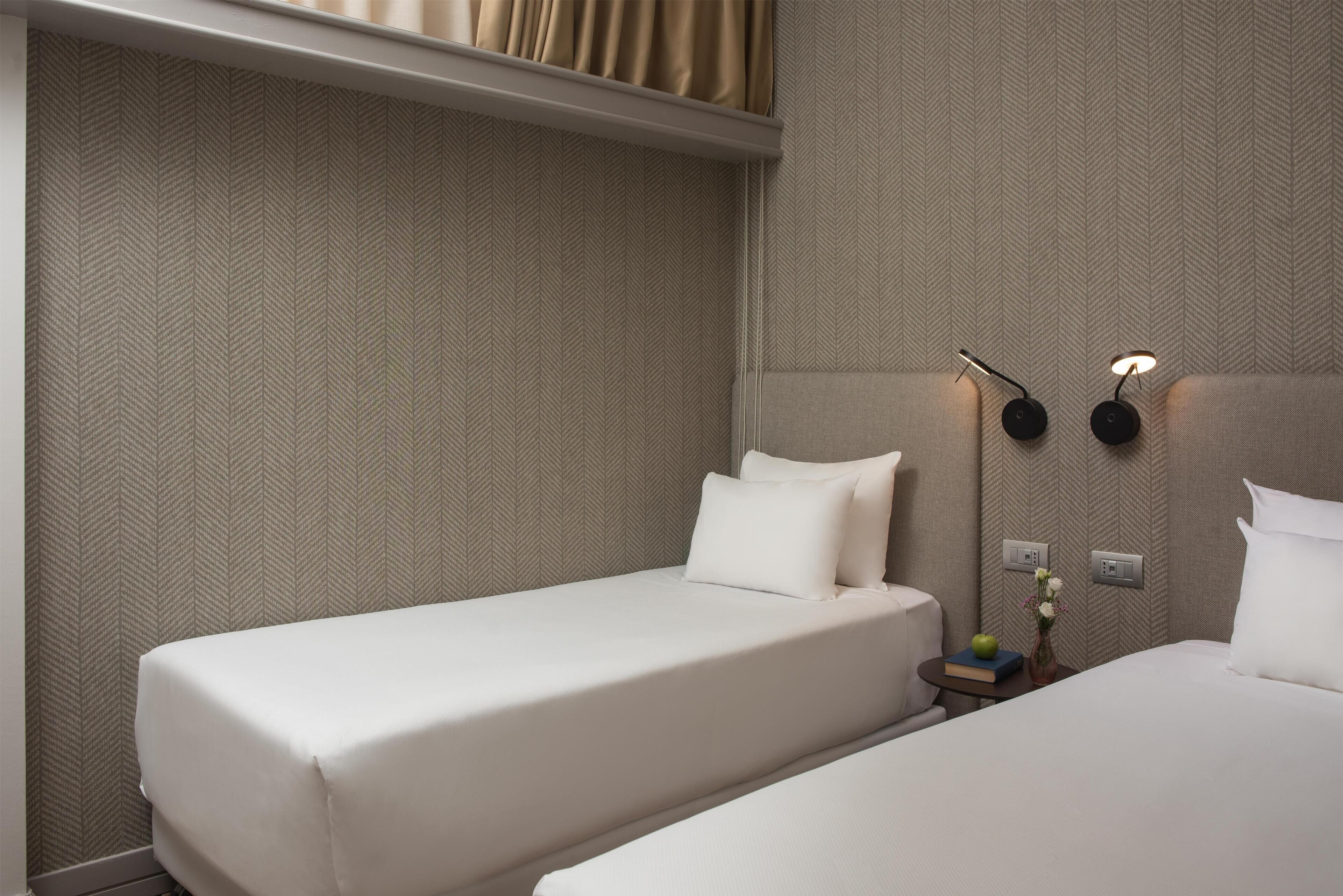 Habitación de hotel con dos camas individuales, una mesita de noche con lámpara y un enchufe en la pared.