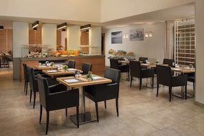A modern dining area with multiple tables and chairs, a central counter with food and beverages, and a large window providing natural light.