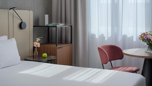 A hotel room with a bed, a small table with a vase of flowers, a chair, a round table with a vase of flowers, a bookshelf, and a window with sheer curtains.