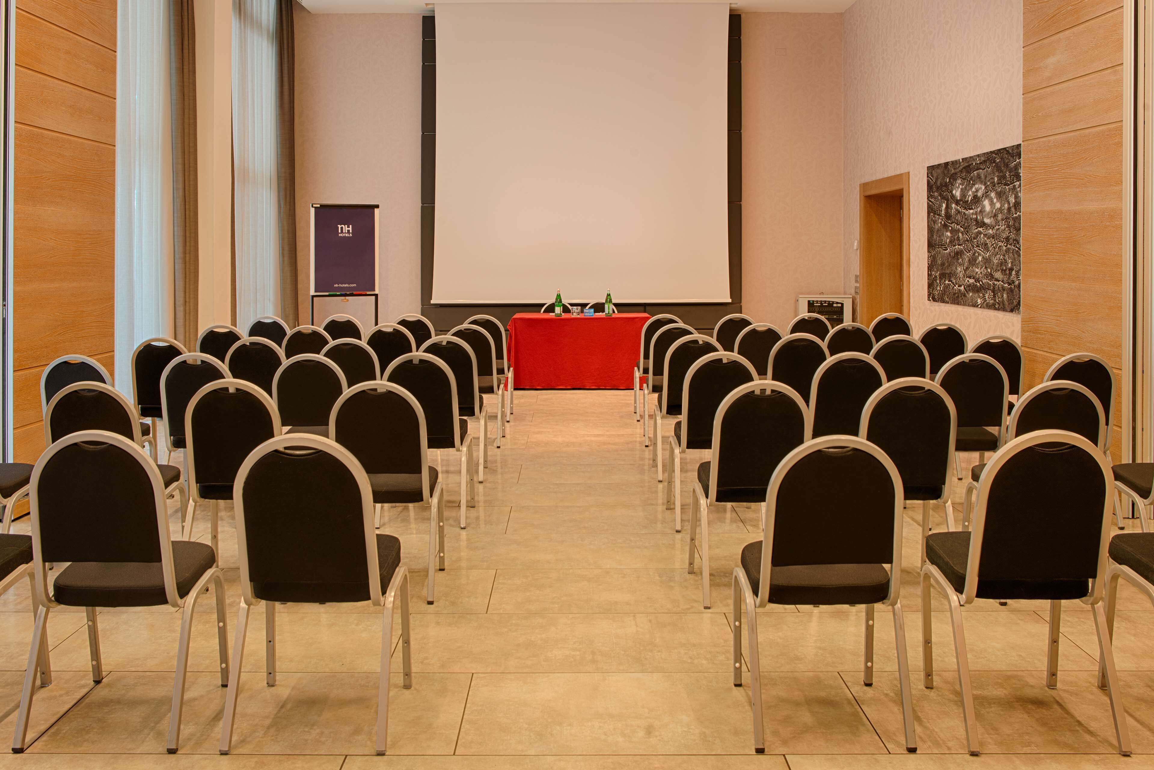 Una sala conferenze con file di sedie rivolte verso un podio con schermo, proiettore e un piccolo tavolo con bottiglie.