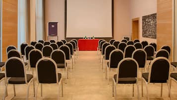 Una sala conferenze con file di sedie rivolte verso un podio con schermo, proiettore e un piccolo tavolo con bottiglie.