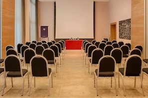 A conference room with rows of chairs facing a podium with a screen, a projector, and a small table with bottles.