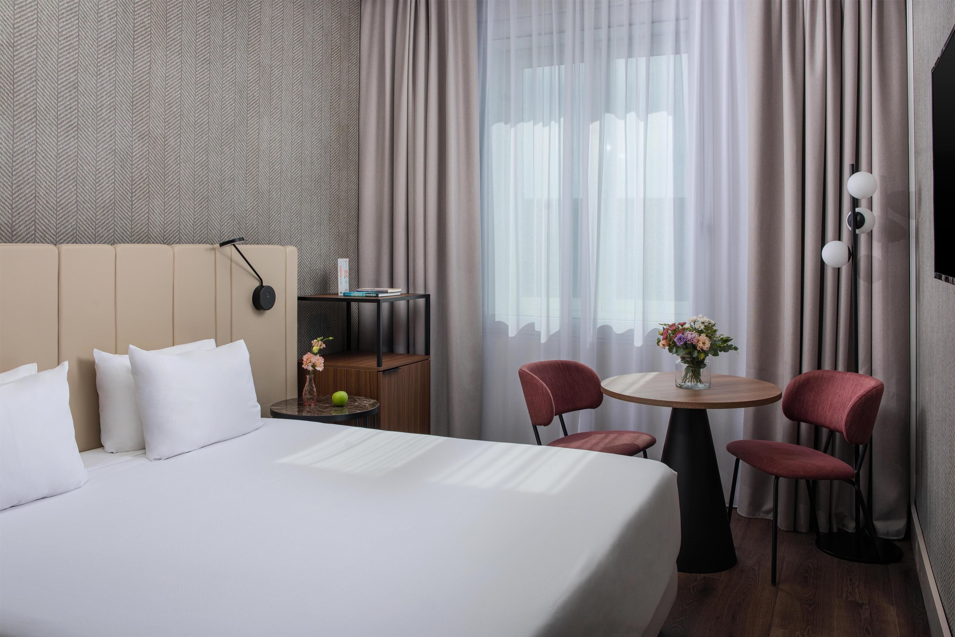 A hotel room with a large bed, a small round table with two chairs, a nightstand with a lamp, and a window with curtains.