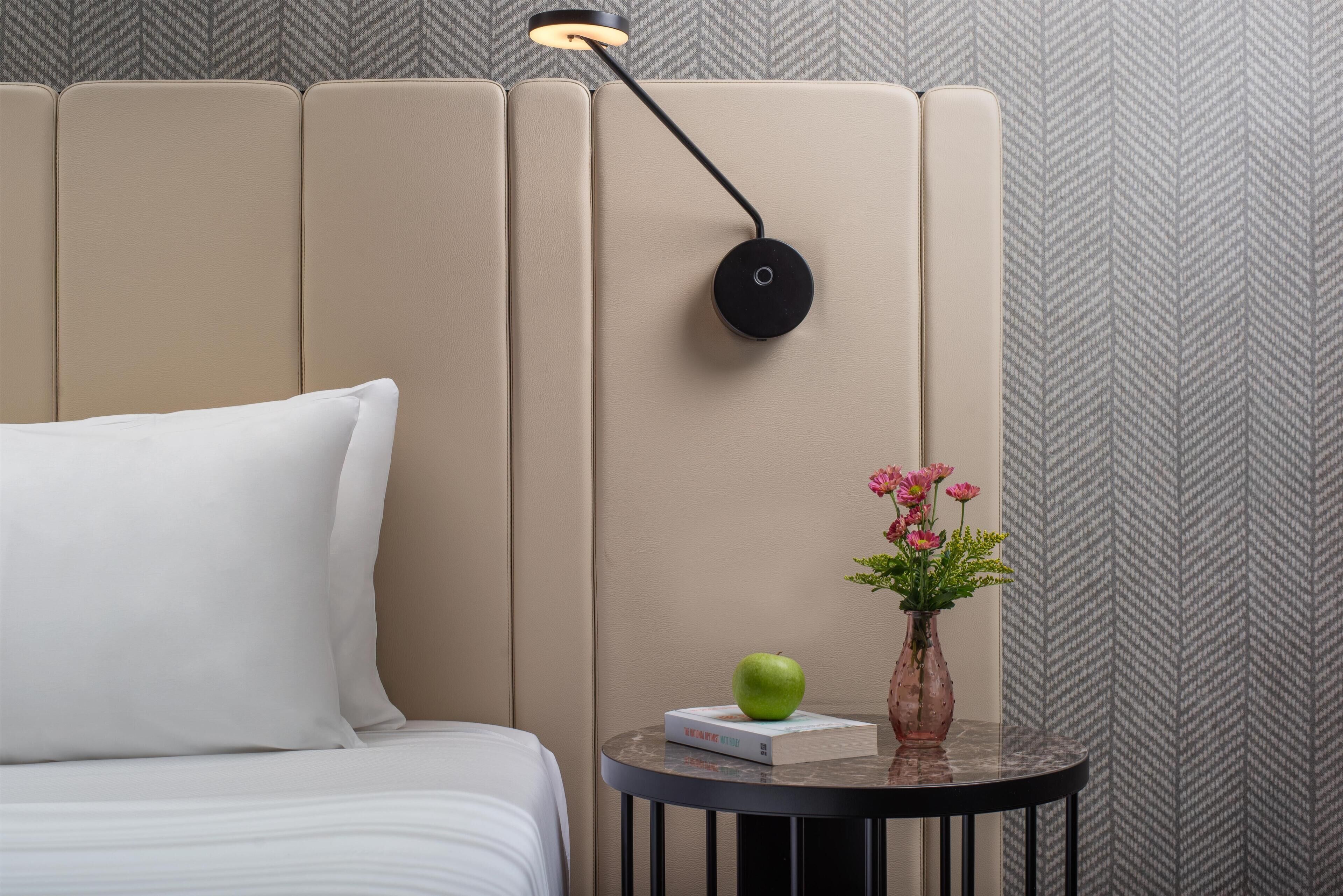 A neatly made bed with a white pillow, a small round table with a book, an apple, and a vase with flowers.