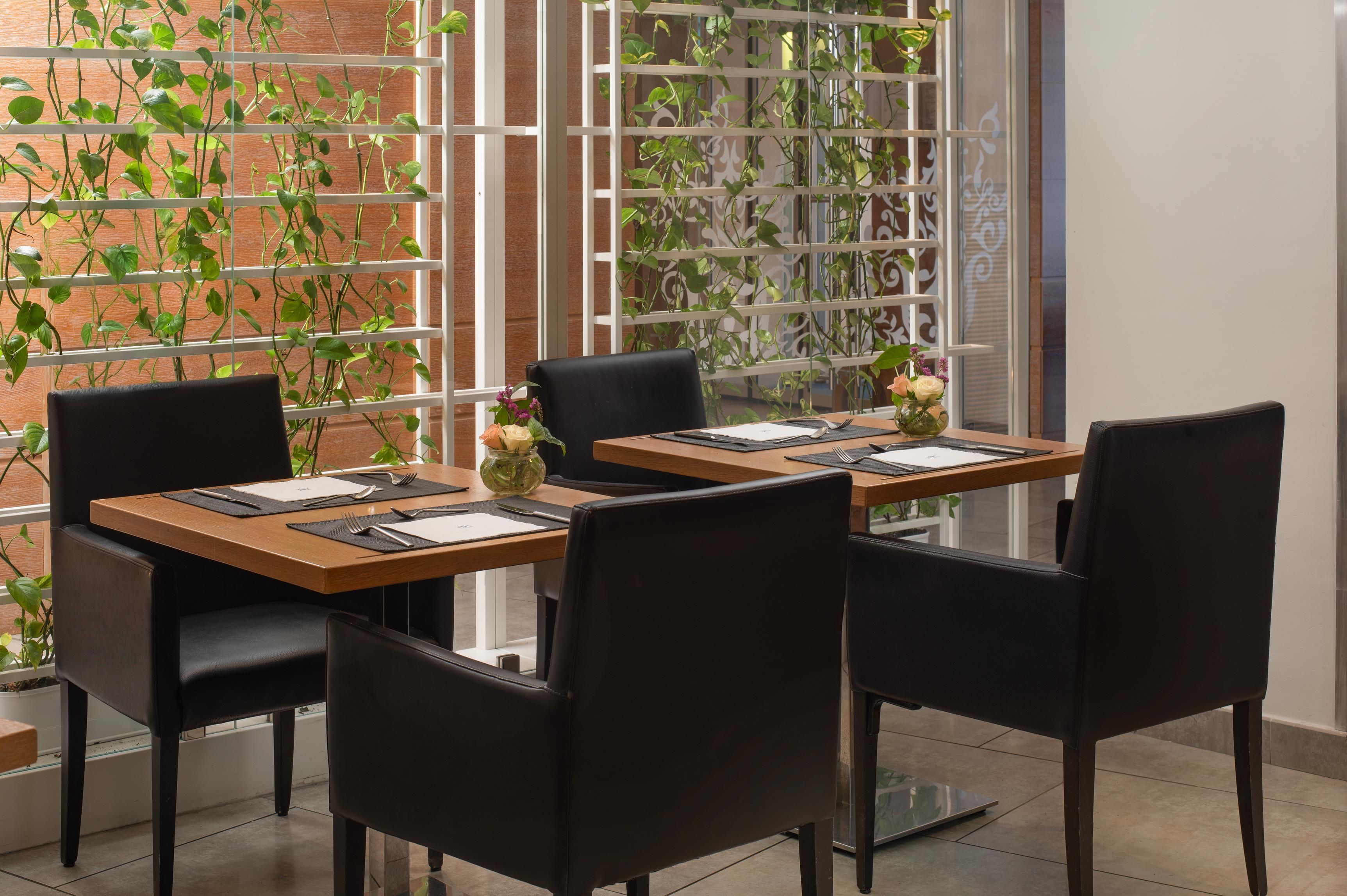 Un coin repas avec une table dressée pour deux personnes, entourée de chaises, et offrant une vue sur un mur de briques et une fenêtre ornée de plantes.