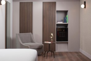 Een hotelkamer met een grijze fauteuil, een klein tafeltje met een vaas, een boekenkast en een spiegel.