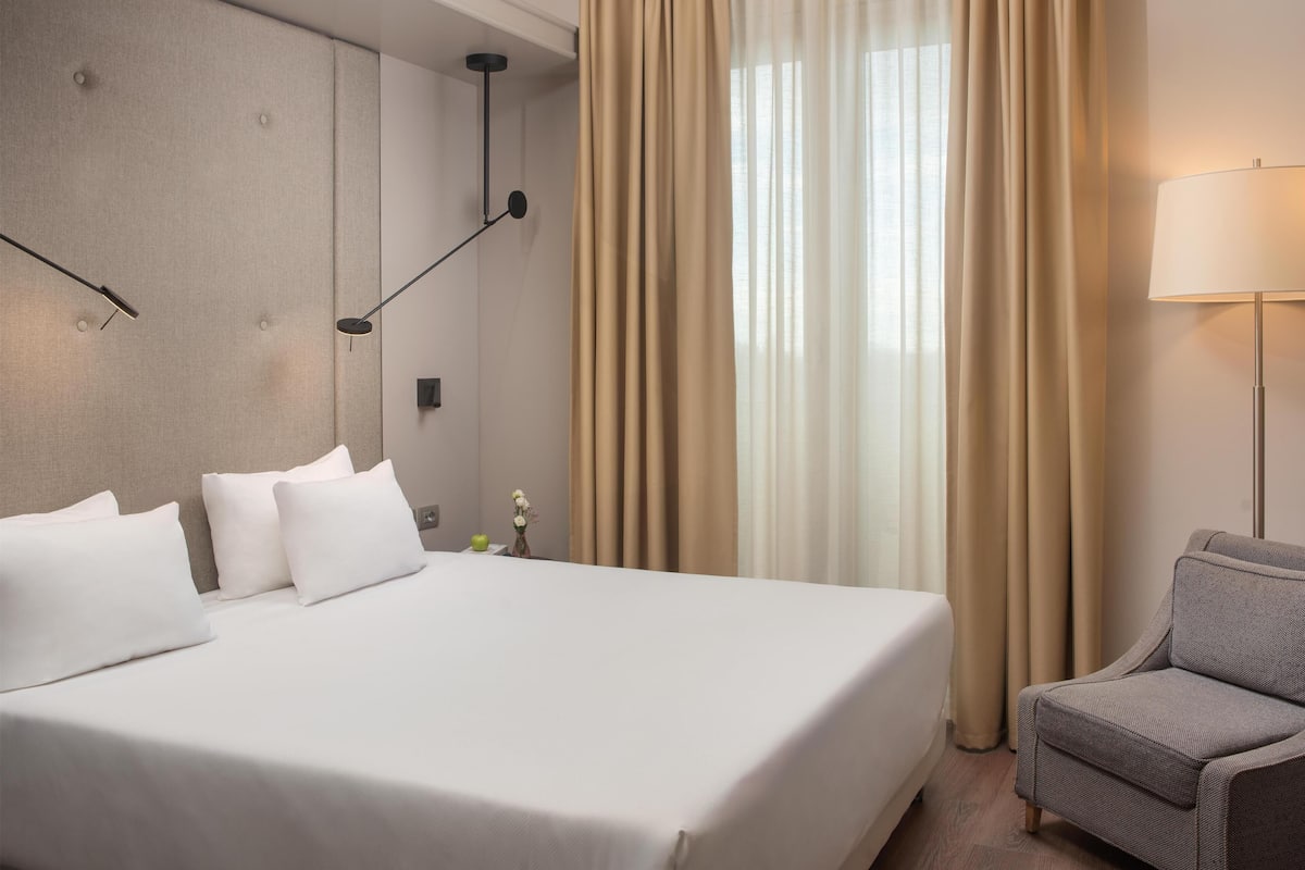 A hotel room with a large bed, a grey armchair, a wall-mounted lamp, and a window with curtains.