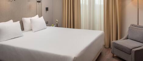Una habitación de hotel con una cama grande, un sillón gris, una lámpara de pared y una ventana con cortinas.