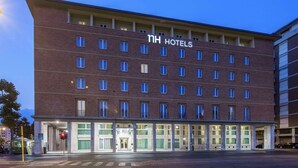 A multi-story hotel with a prominent "NH Hotels" sign, featuring a modern brick facade and a ground-level entrance with glass doors.