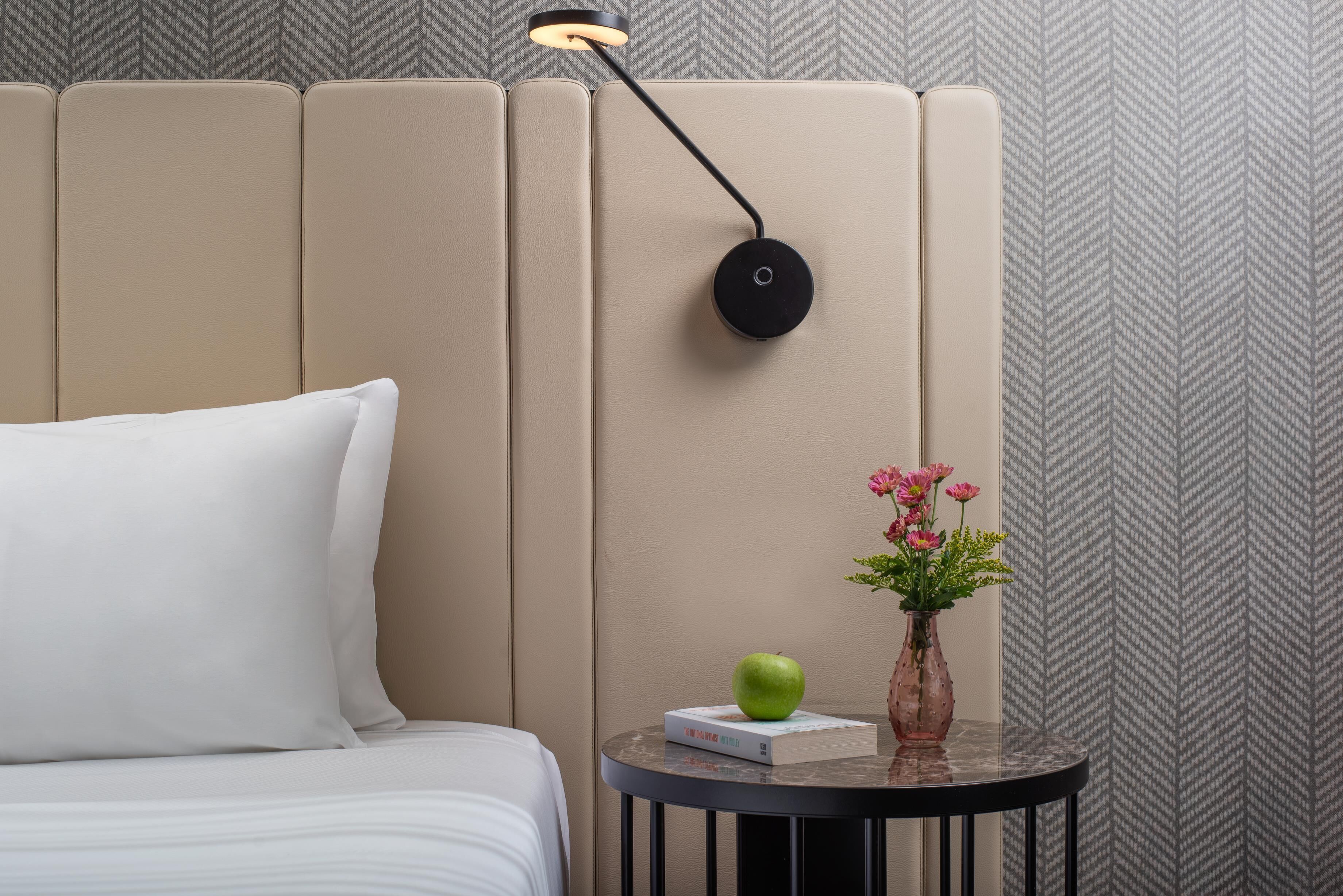 Un dormitorio con una cama, una mesita de noche con un libro, una manzana, un jarrón con flores y una lámpara.