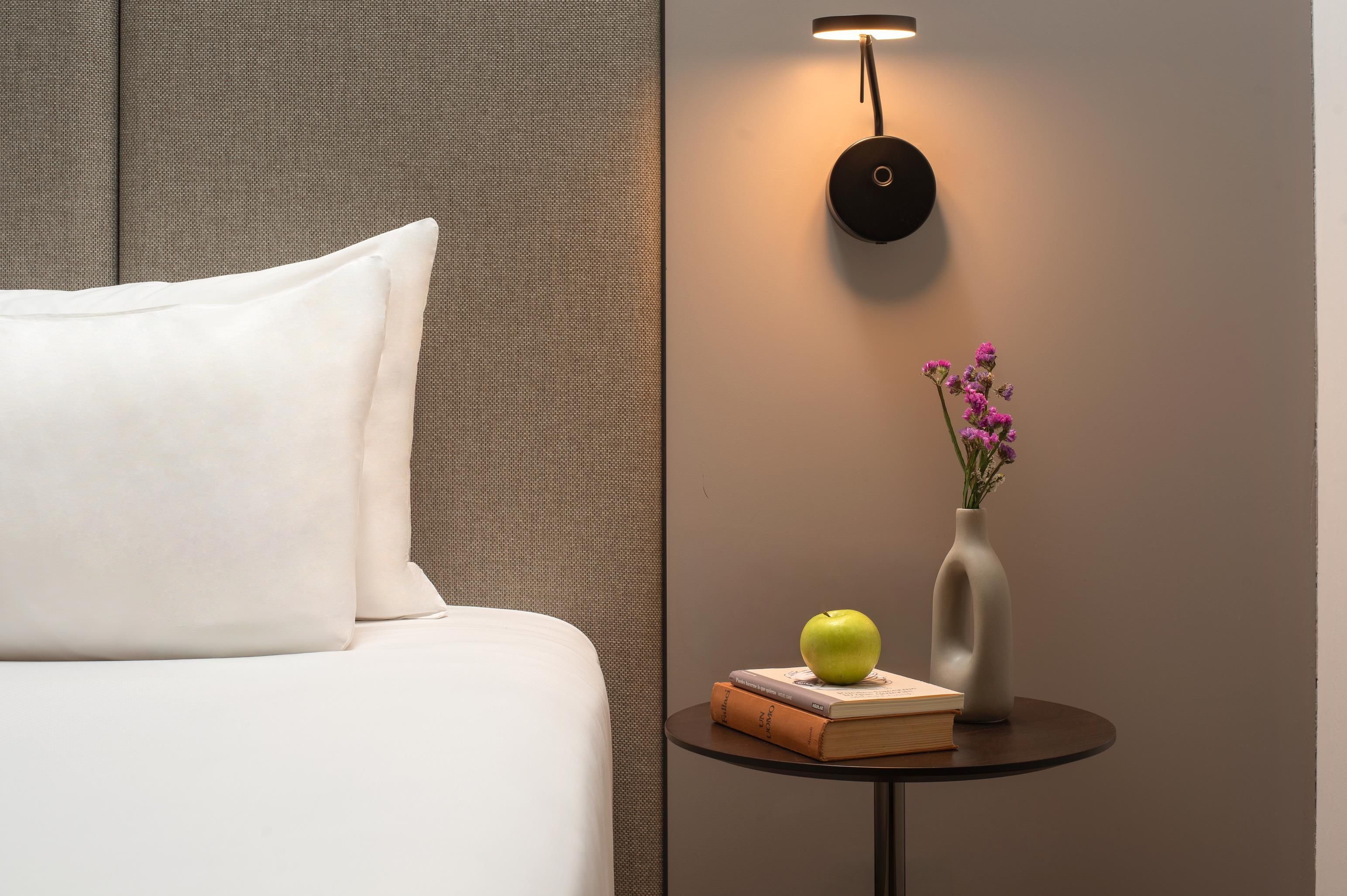 Un lit bien fait, avec un oreiller blanc, une petite table sur laquelle se trouve un vase de fleurs, une pomme et un livre.