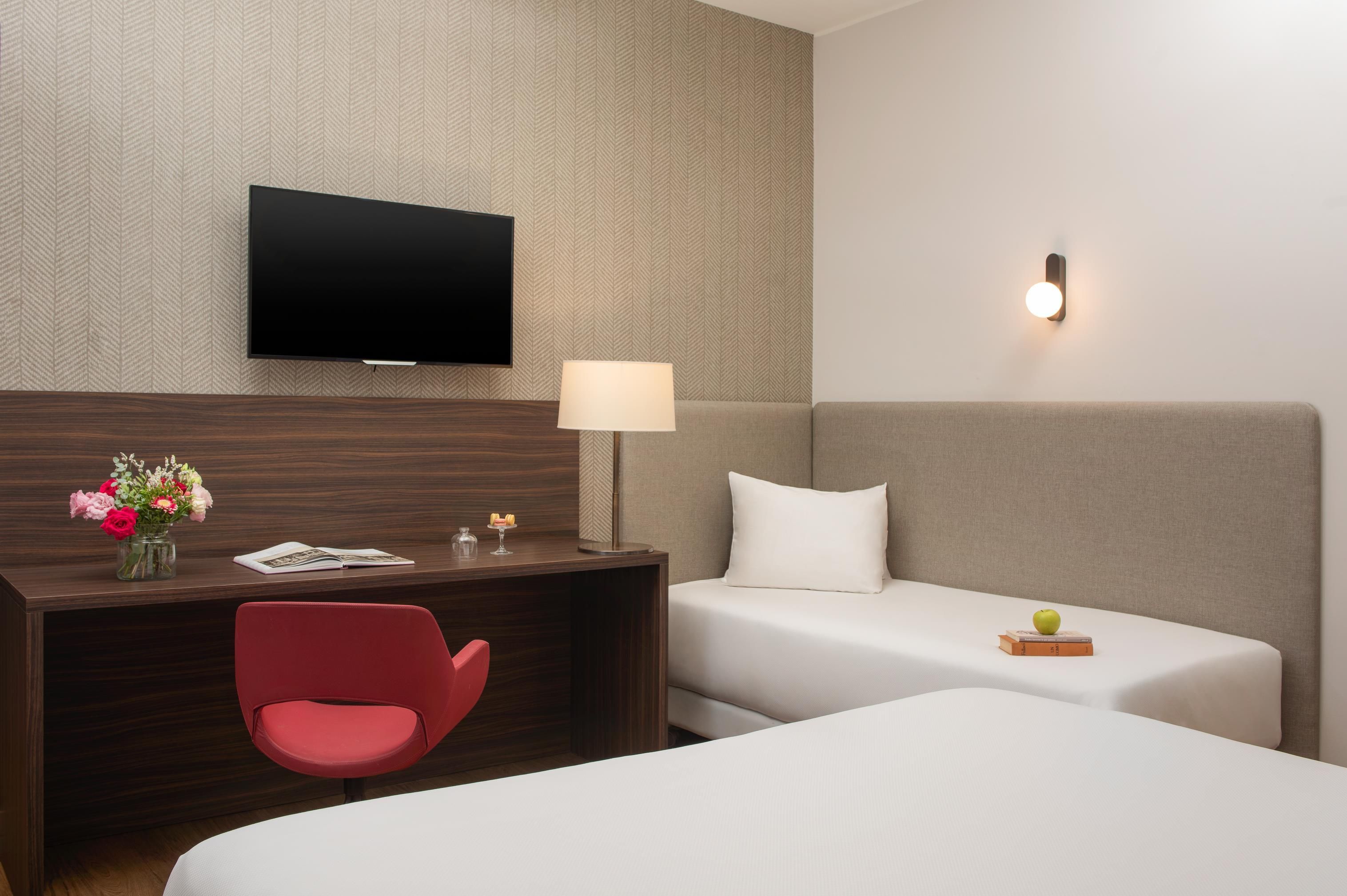 A hotel room with a flat-screen TV mounted on the wall, a desk with a lamp, a red chair, two beds with white linens, and a small table with a vase of flowers.