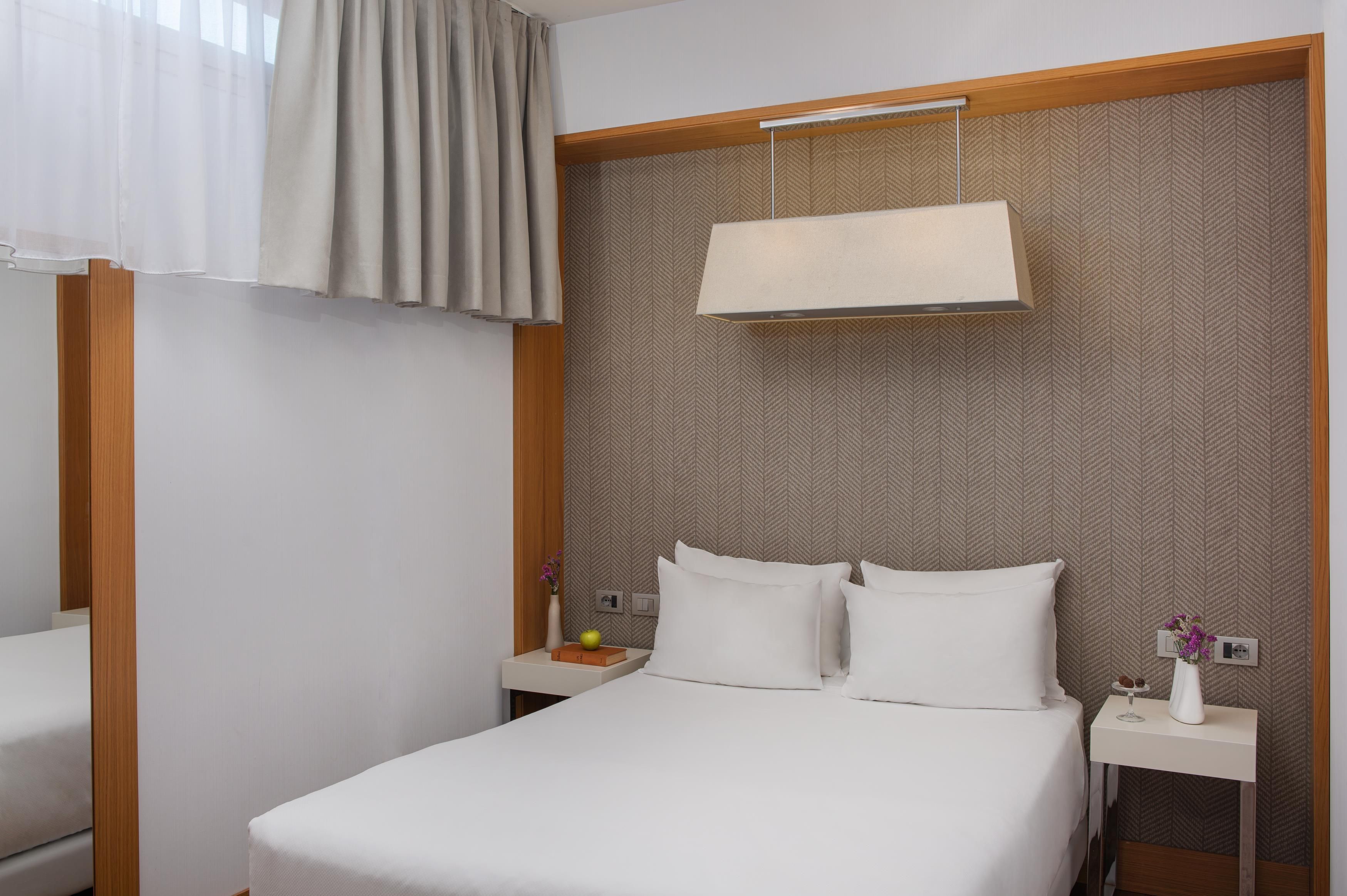 A hotel room with a bed, two nightstands, a lamp, a vase with flowers, and a wall with a herringbone pattern.