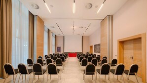 A conference room with rows of chairs facing a stage, large windows, and a modern ceiling design.