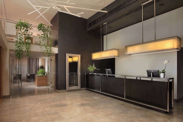 A modern reception area with a dark reception desk, a glass partition, and a seating area with a plant and a potted orchid. - NH Pisa (Pisa)