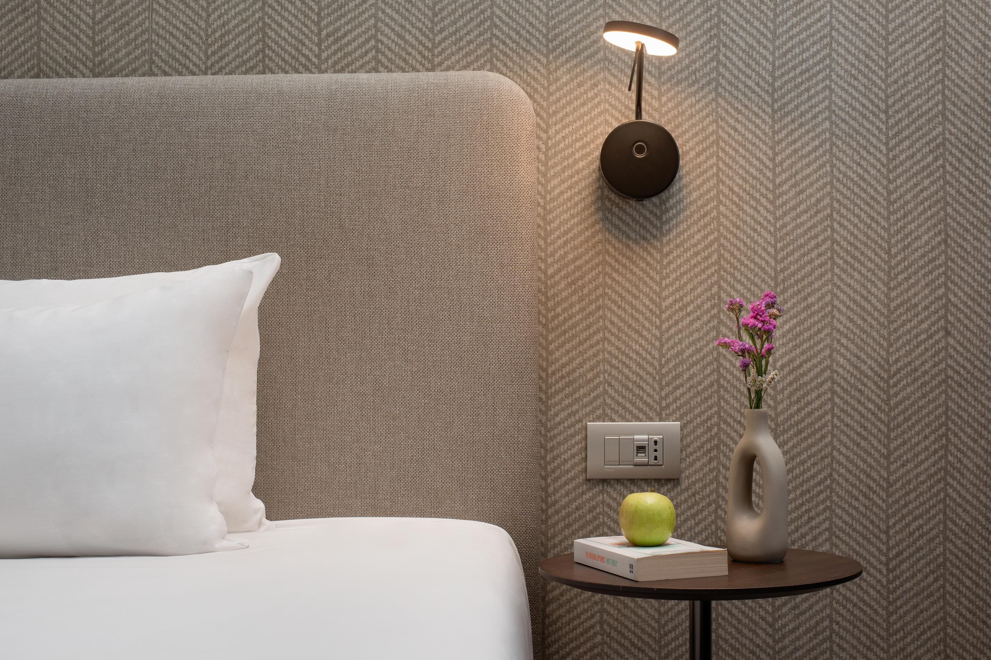 Una cama bien hecha con un almohadón blanco, una mesita con un libro y una manzana, un florero con flores, y una lámpara de pared.