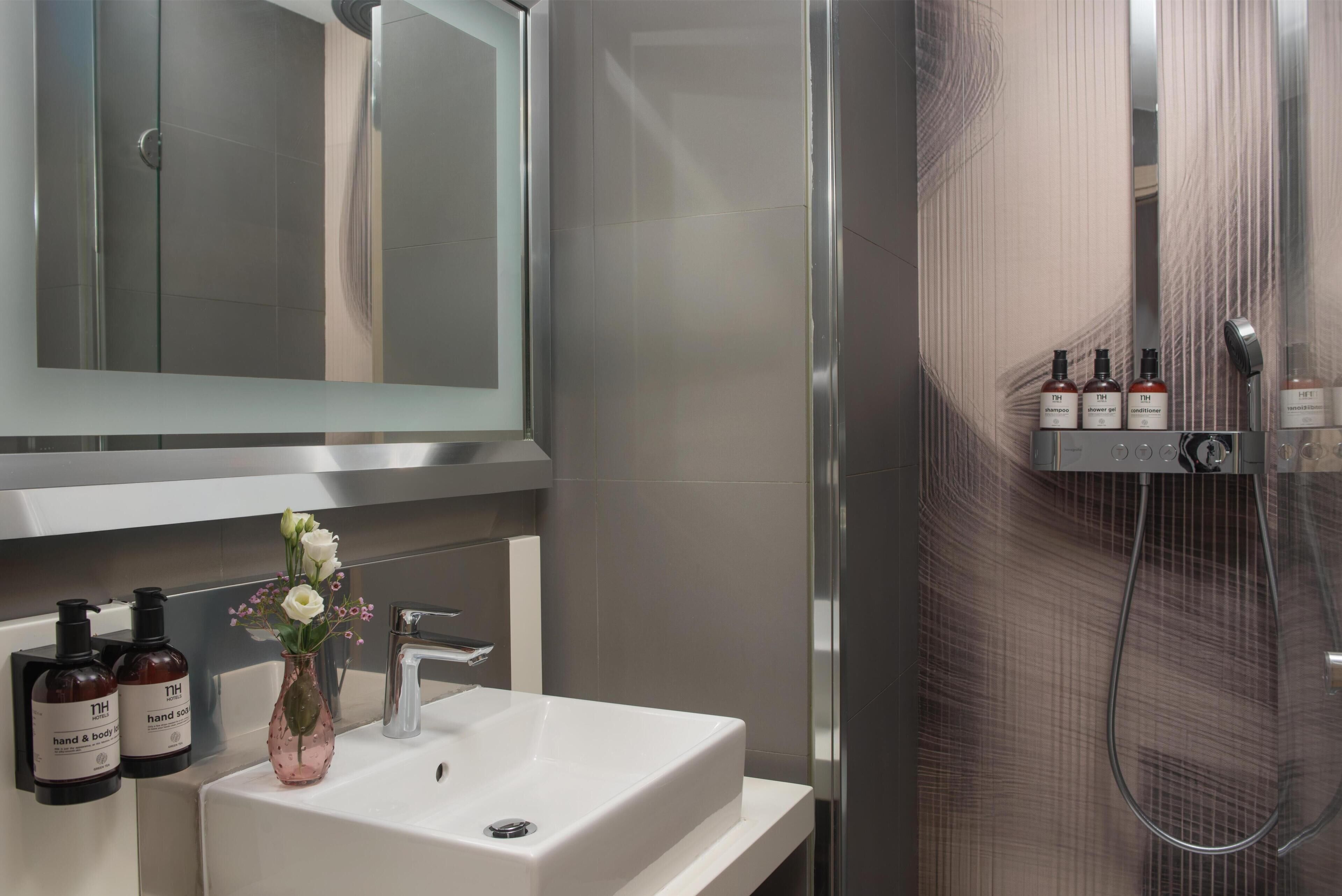 Une salle de bain moderne avec un lavabo blanc, un miroir et un espace douche. On trouve des articles de toilette sur le lavabo et une pomme de douche au-dessus de la douche.