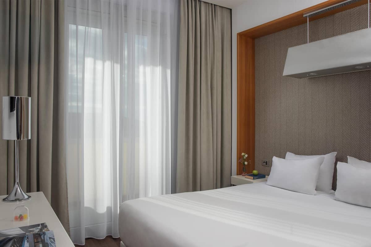 A hotel room with a large bed, a bedside table with a lamp, a nightstand with a vase and books, and sheer curtains.
