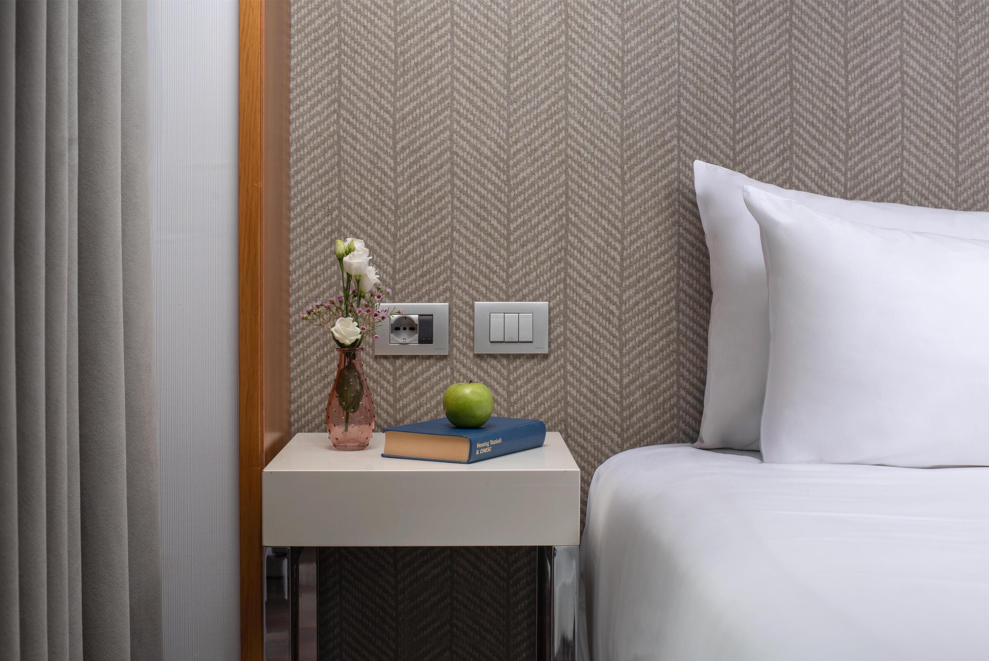 A neatly made bed with white bedding, a bedside table with a book, an apple, and a vase with flowers.