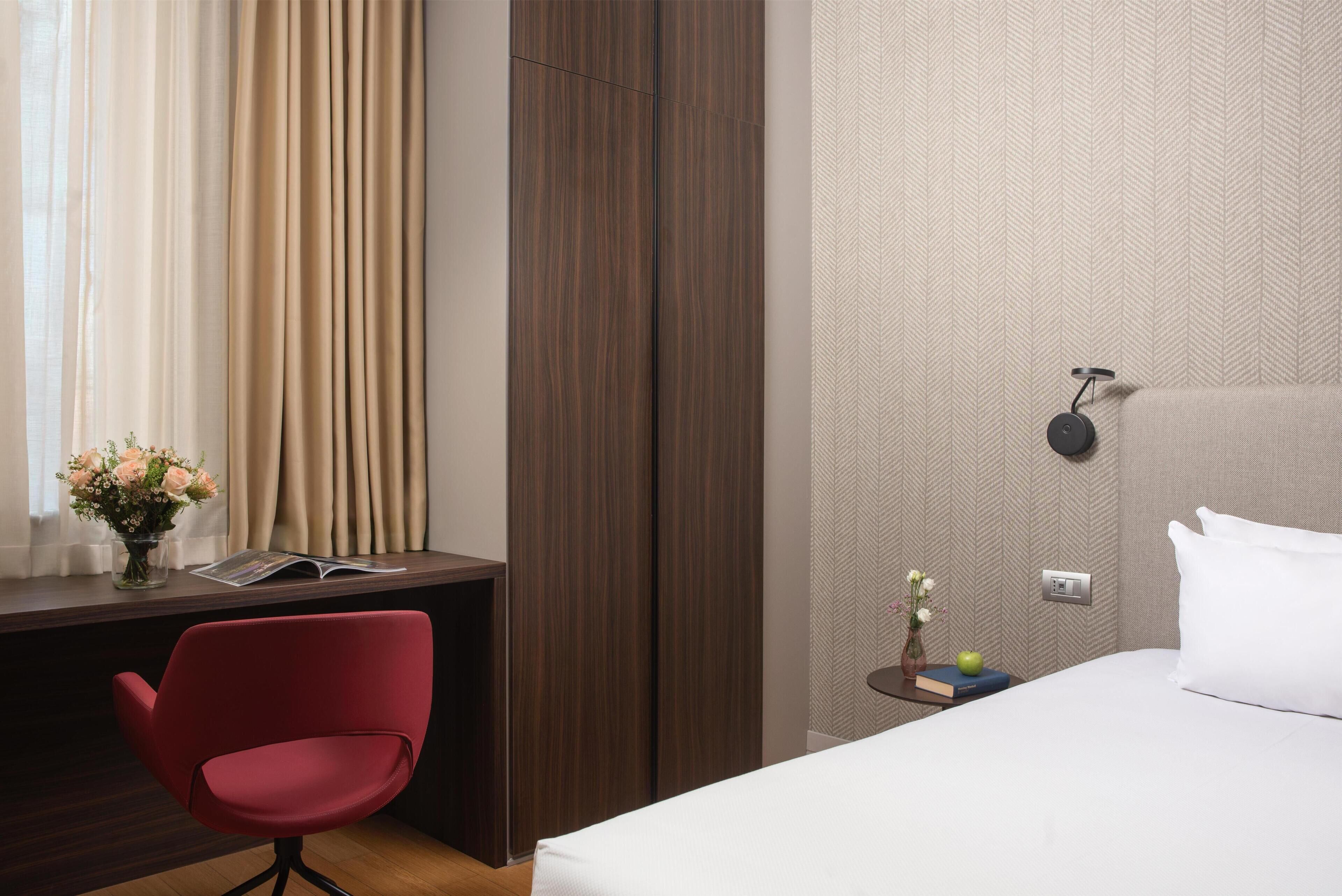 A hotel room with a bed, a desk with a chair, a vase with flowers, a book, and a nightstand with a lamp.