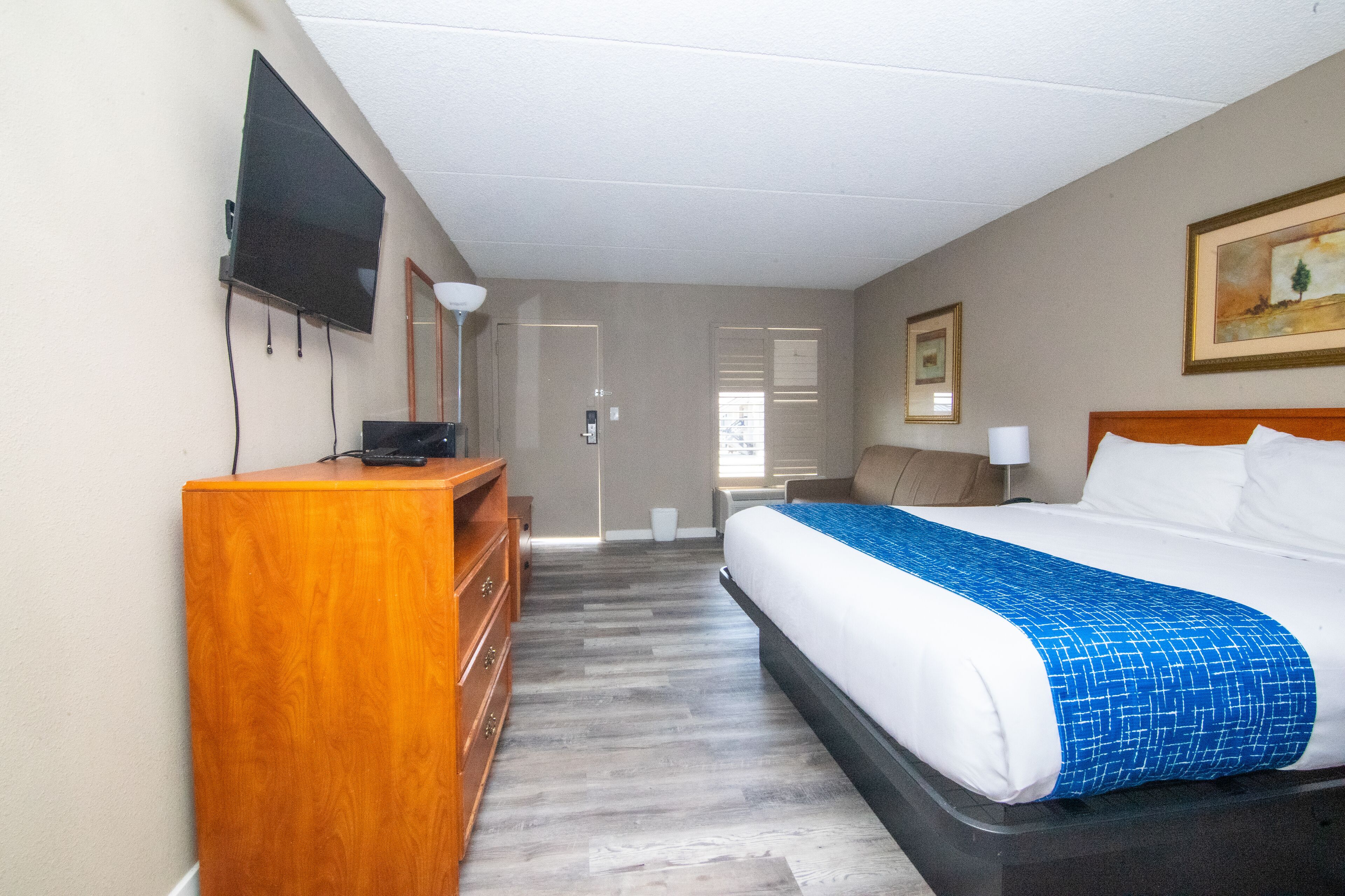 A hotel room with a bed, a wooden dresser, a flat-screen TV mounted on the wall, and a painting on the wall.