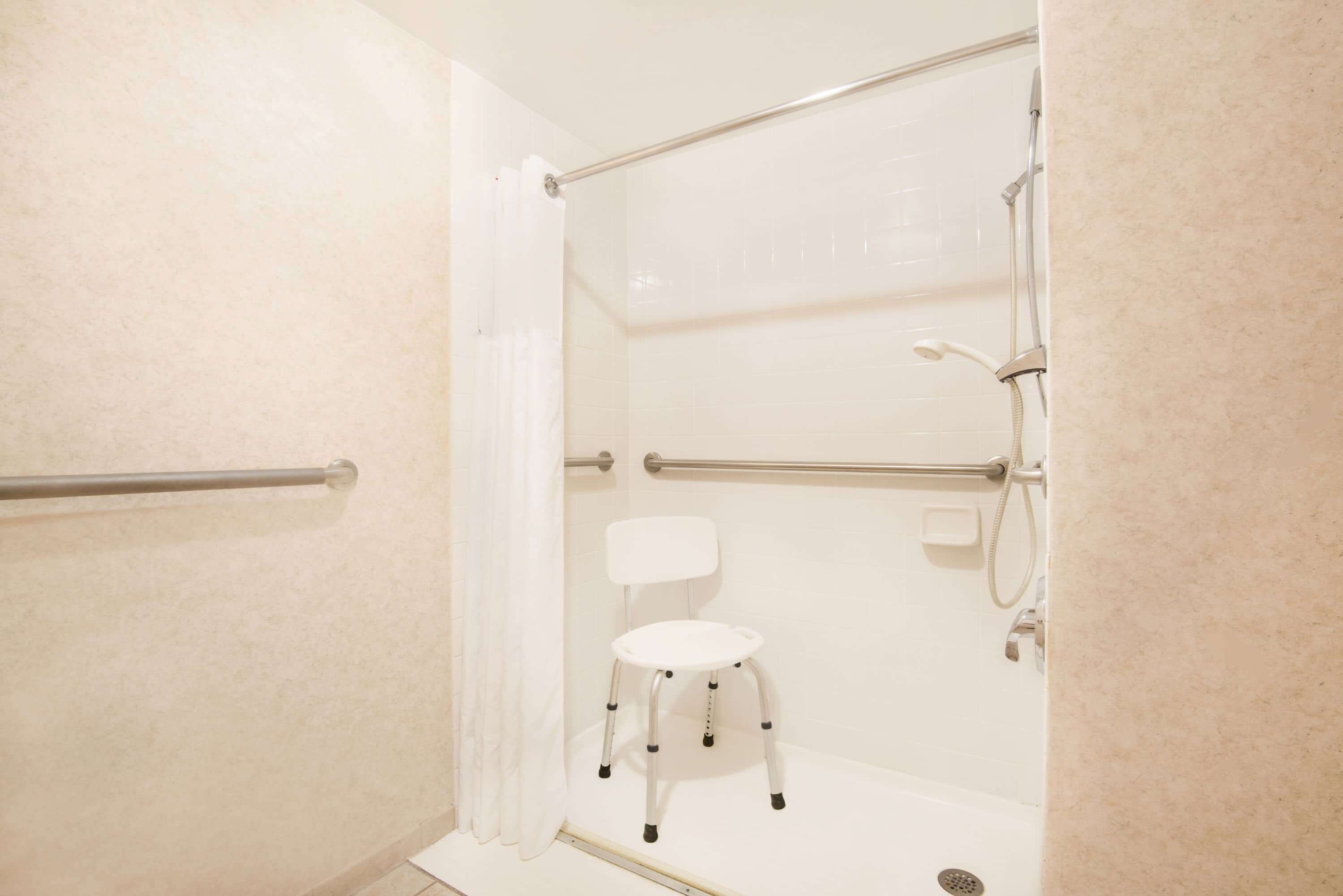 A shower area with a white chair, grab bars, and a handheld showerhead.