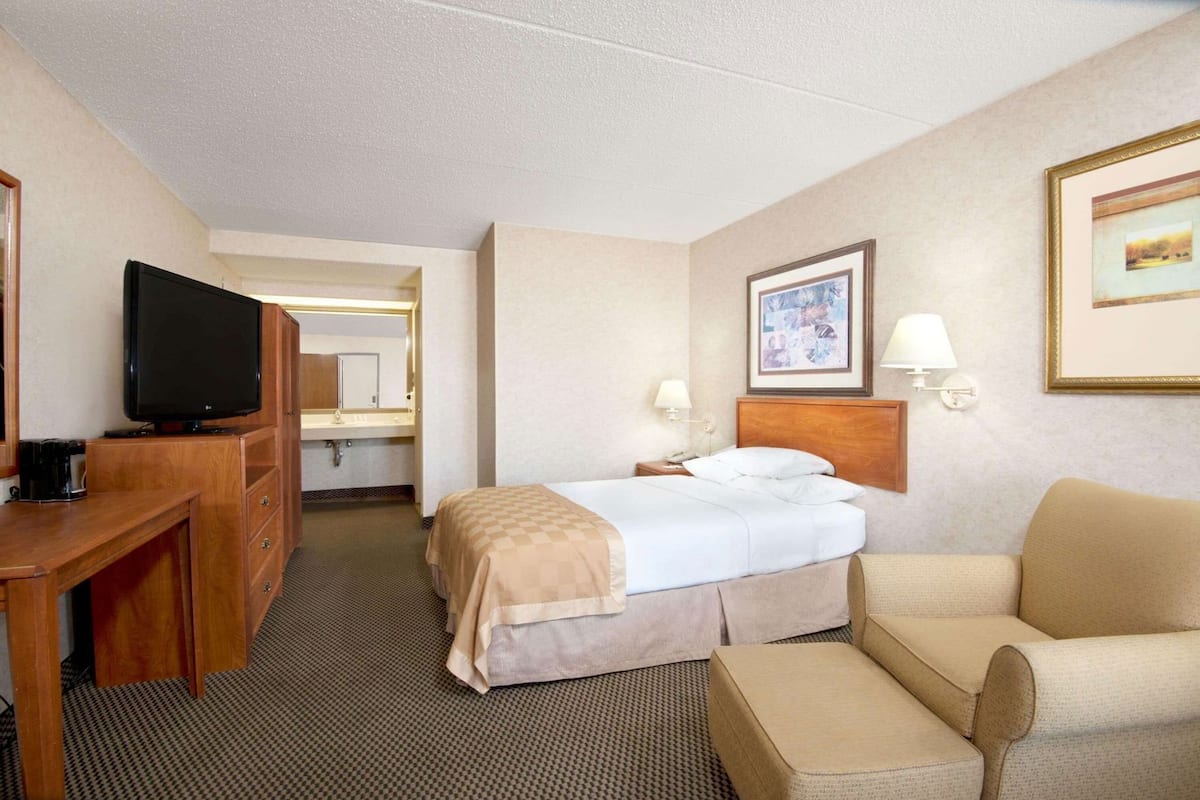 A hotel room with a bed, a desk with a TV, a chair, a lamp, and a framed picture on the wall.