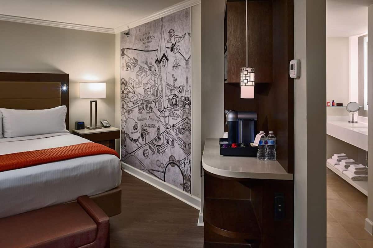 A hotel room with a bed, a desk with a coffee maker, a map on the wall, and a bathroom with a mirror and towel rack.