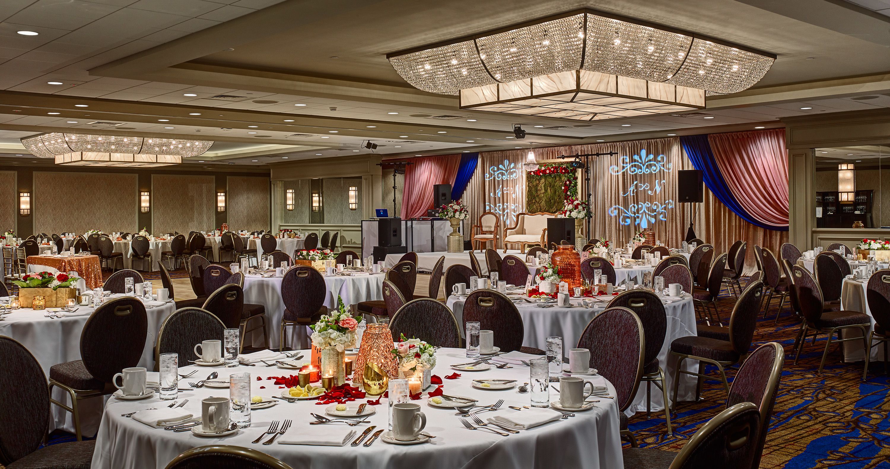 Un salón de banquetes con mesas redondas dispuestas para comer, un escenario con un telón de fondo y una araña.