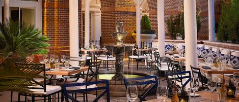 An outdoor dining area with brick walls, white columns, and string lights. Tables are set with wine glasses and plates.