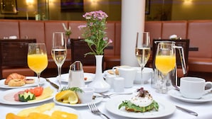 Une table mise pour le petit-déjeuner, avec des assiettes remplies de nourriture, des verres de jus d’orange et de vin blanc, et un vase de fleurs.