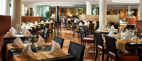 Un restaurant spacieux avec de nombreuses tables, toutes dressées avec des serviettes blanches, des verres et des couverts. On y trouve plusieurs chaises à haut dossier, ainsi qu’un mélange de banquettes et de tables. Le plafond est doté d’un éclairage encastré et présente un design incurvé.