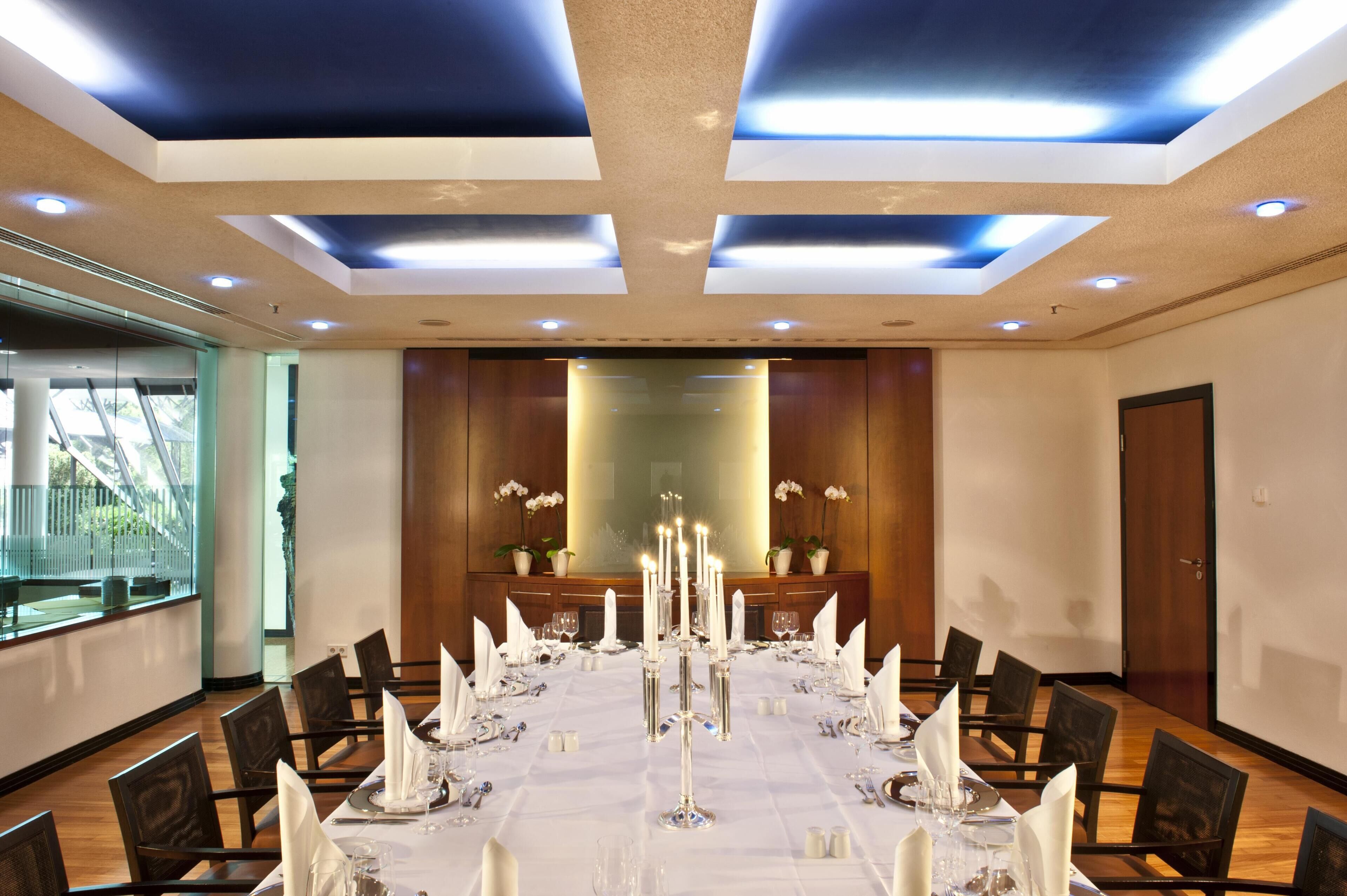 Un salón de conferencias con una larga mesa preparada para una reunión, equipado con iluminación moderna, paneles de madera y un espejo grande.