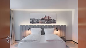 Une chambre d’hôtel moderne avec un grand lit, un tableau représentant un paysage urbain au-dessus du lit et une vue sur un château.